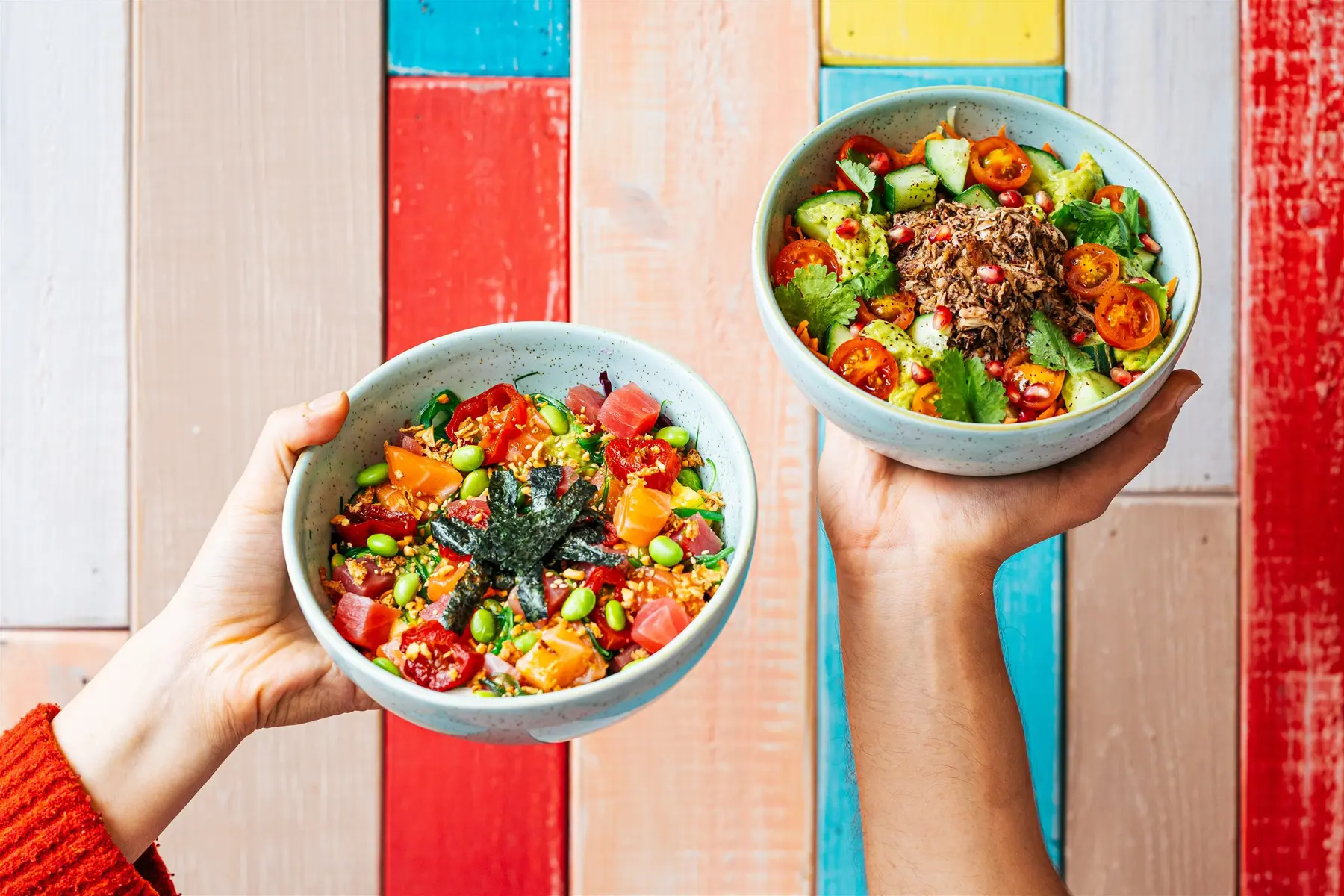 Two hands holding Peeld bowls over a colourful painted wooden table: the All Out poke bowl and the chicken Shawarma bowl