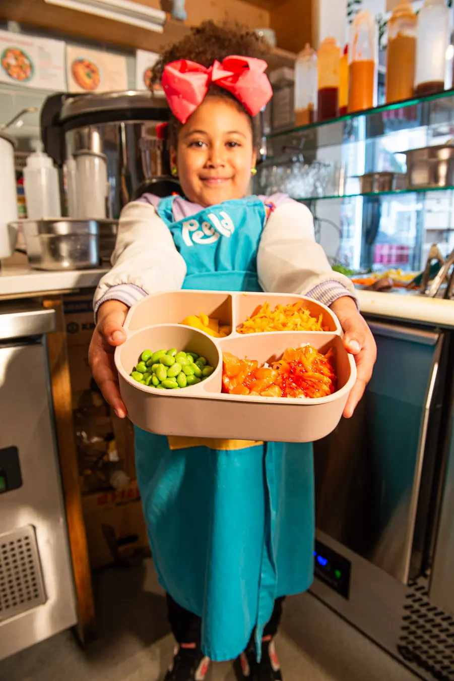 A young customer in a Peeld apron holding a kids' bento box of edamame, mango, carrot and salmon