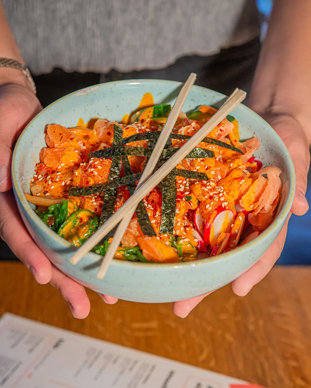 A salmon poke bowl with sriracha mayo, nori, pickled ginger and radish, held over a table at Peeld
